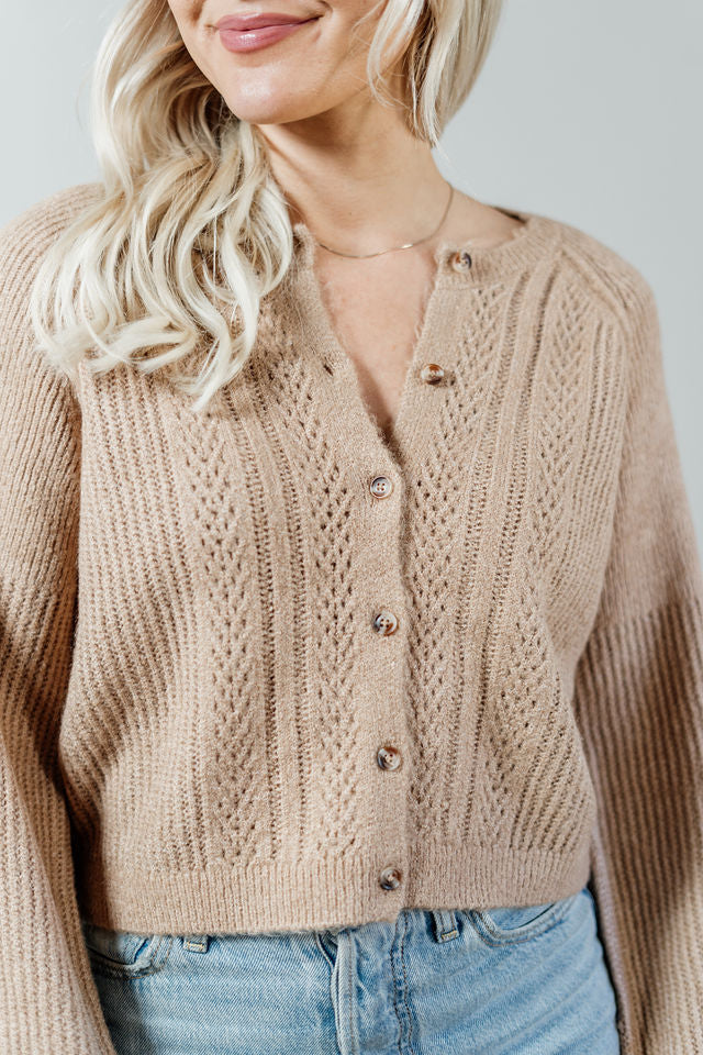 Pictured is a beige, short cardigan with a button-down front, cuffed sleeves, and grandma-knit design.