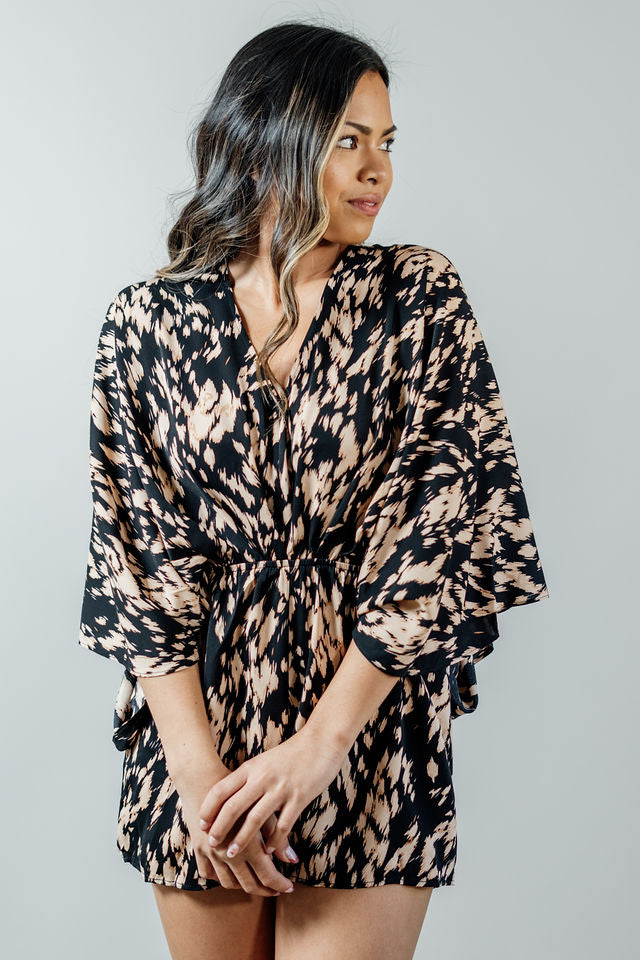 Pictured is a black long sleeve romper with a tan abstract pattern, ruched front, cinched waist, and flowy bottoms.
