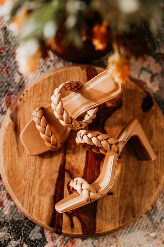 Pictured is a tan, heeled sandal with two braided straps and thin heel.