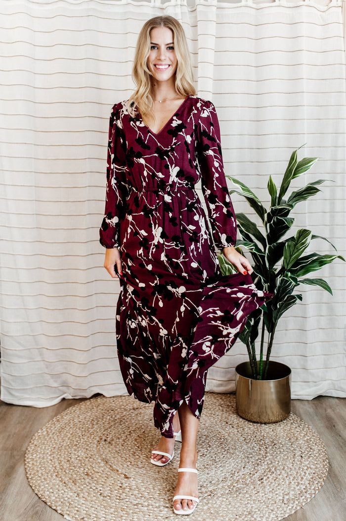 Pictured is a burgundy, long sleeve maxi dress with a lace trim v-neckline, puffed long sleeves, cinched waist, and side slit.
