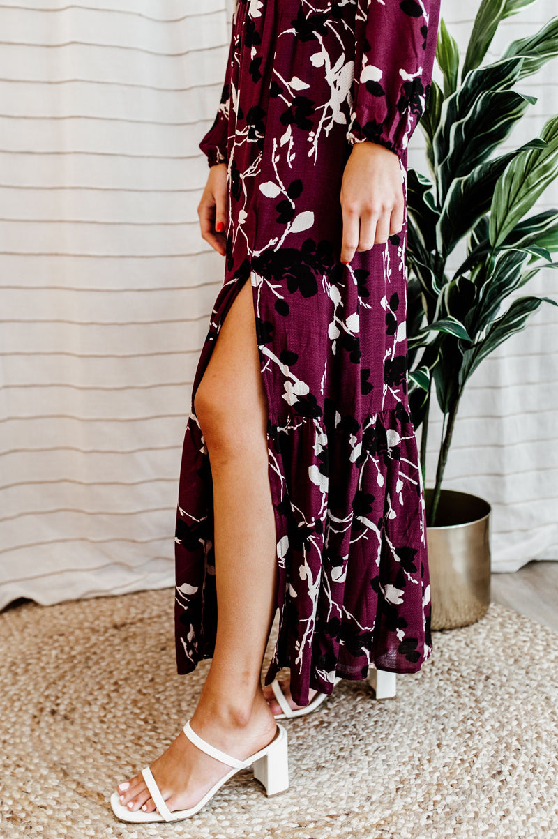 Pictured is a burgundy, long sleeve maxi dress with a lace trim v-neckline, puffed long sleeves, cinched waist, and side slit. 