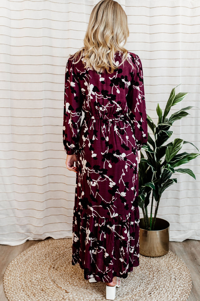 Pictured is a burgundy, long sleeve maxi dress with a lace trim v-neckline, puffed long sleeves, cinched waist, and side slit. 