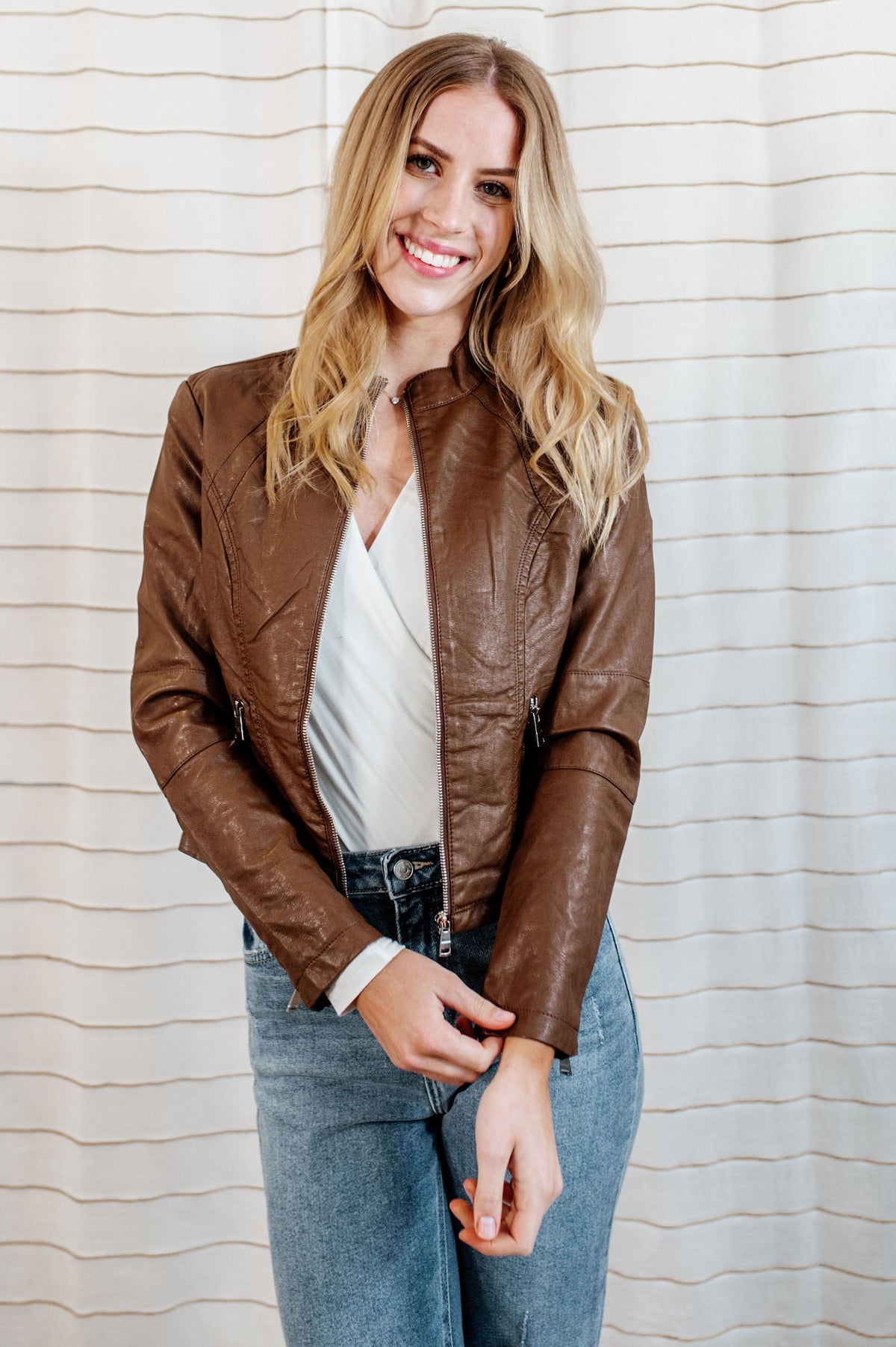 Pictured is a chocolate-brown, vegan faux leather jacket with an outside stitch detailing, floral-lined inside, and zipper detailing on the front, sleeves, and pockets.