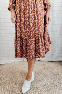 Pictured is a burnt-orange, long sleeve midi dress with a v-neckline, wrap style bust, ruffled waistline and detailing, and flowy long sleeves. 