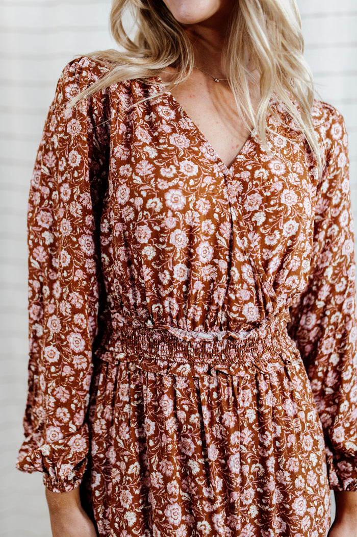 Pictured is a burnt-orange, long sleeve midi dress with a v-neckline, wrap style bust, ruffled waistline and detailing, and flowy long sleeves.