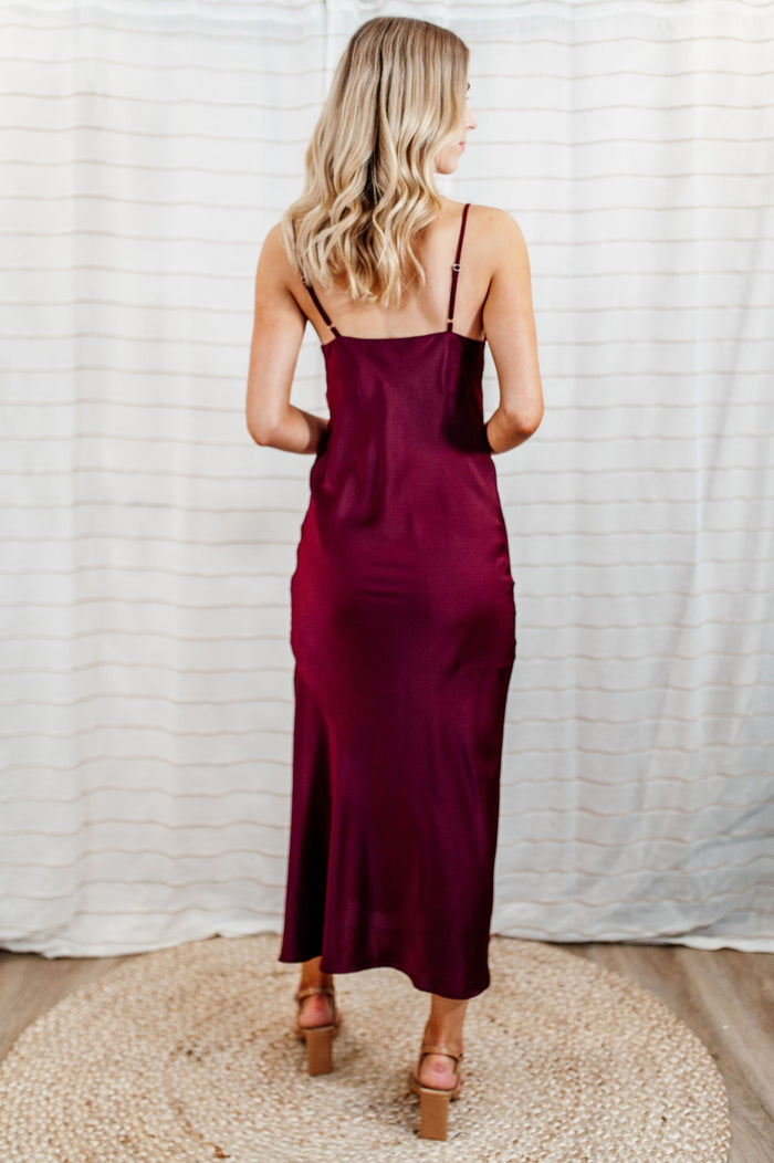 Pictured is a red, stunning and silky satin slip dress with a flattering cowl neckline, slit on the side, adjustable straps, and midi length.