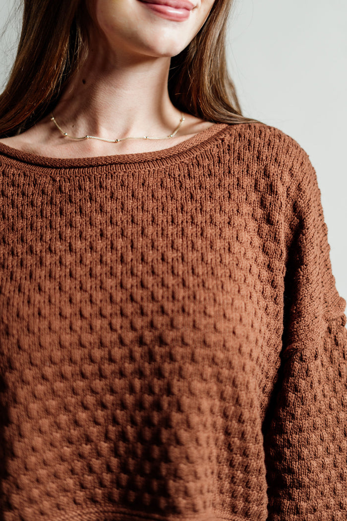 Pictured is a red-brown, knit sweater with a scoop neckline, cropped body, and cuffed knit sleeves.