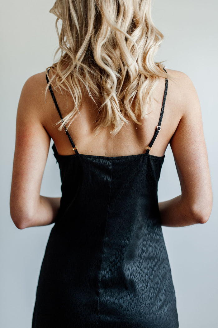 Pictured is a black satin dress with a v-neckline, adjustable straps, and subtle animal print.