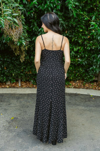 Woman wearing a black polka dot dress standing in front of green foliage.