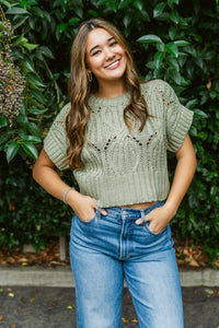 Woman wearing a green sweater and blue jeans standing in front of a green bush