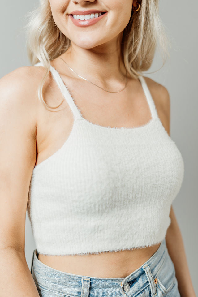 Pictured is a white, knit top with fuzzy material, thin shoulder straps, and cropped body. 
