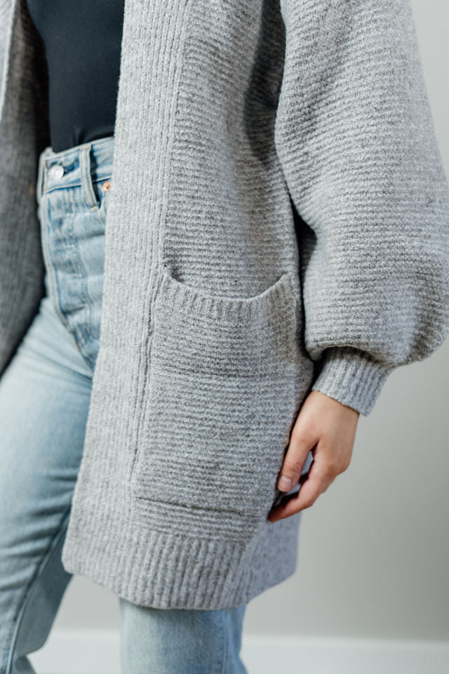 Pictured is a light grey, thick cardigan with balloon sleeves, long body, open front, and front pockets.