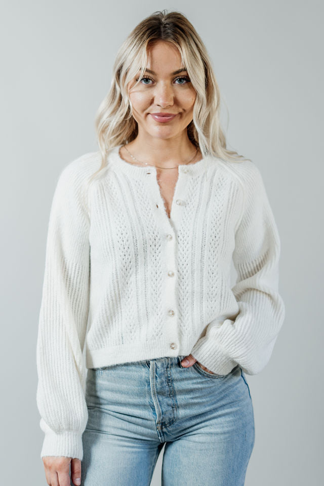 Pictured is a white, short cardigan with a button-down front, cuffed sleeves, and grandma-knit design.