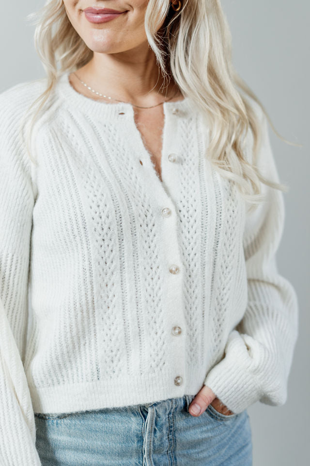 Pictured is a white, short cardigan with a button-down front, cuffed sleeves, and grandma-knit design.