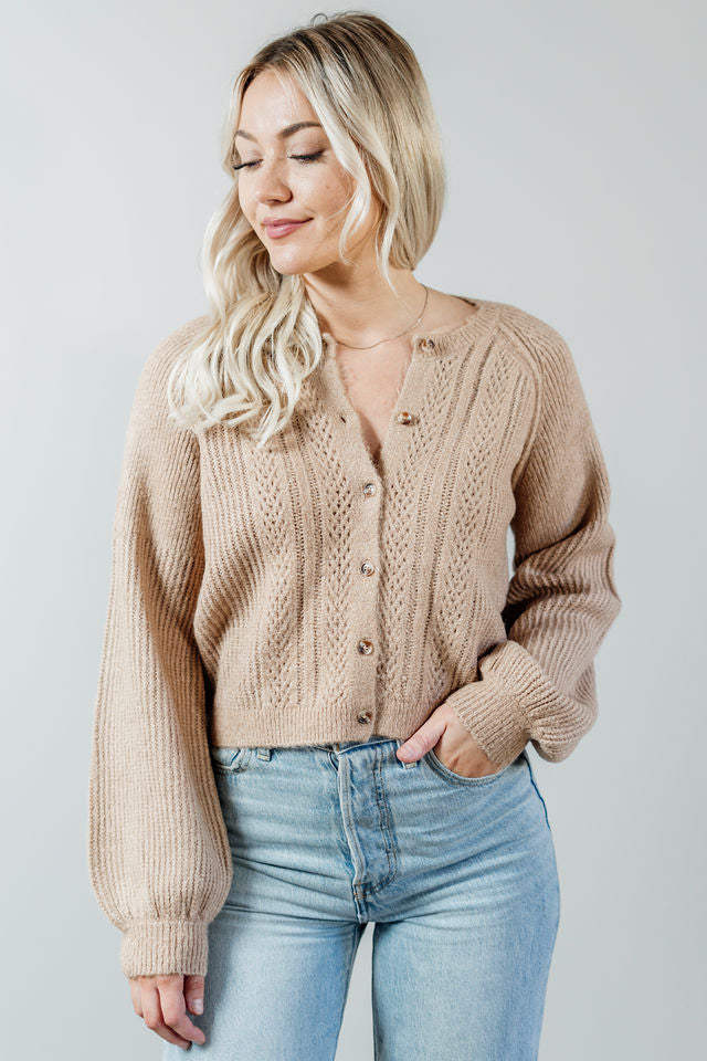 Pictured is a beige, short cardigan with a button-down front, cuffed sleeves, and grandma-knit design.