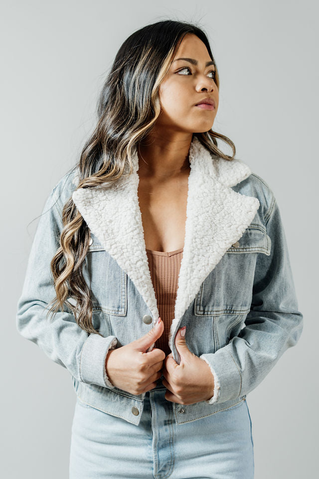 Pictured is a light-wash jean jacket with a white fuzzy collar, cuffed sleeves, button-down front, and elastic waistband.