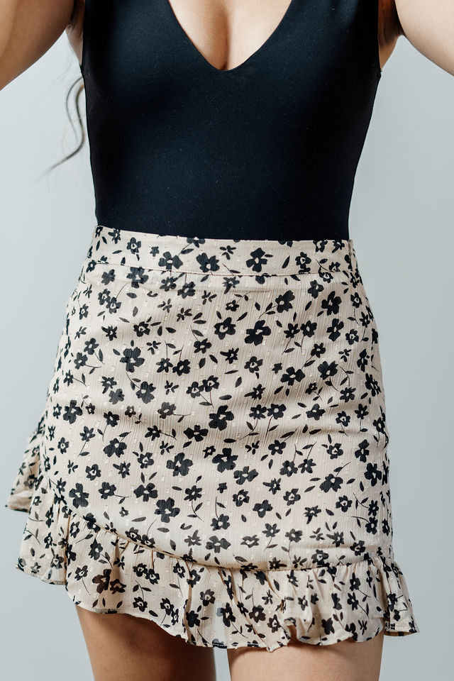 Pictured is a beige mini skirt with a black delicate floral pattern, structured body, seamless zipper, and ruffled hem.