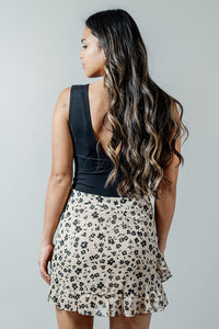 Pictured is a beige mini skirt with a black delicate floral pattern, structured body, seamless zipper, and ruffled hem.