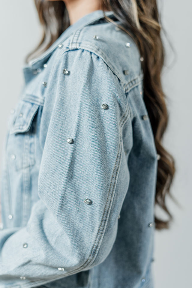Pictured is a classic, blue denim jacket with a collar, button-down front, cuffed sleeves, and diamond detailing all over.