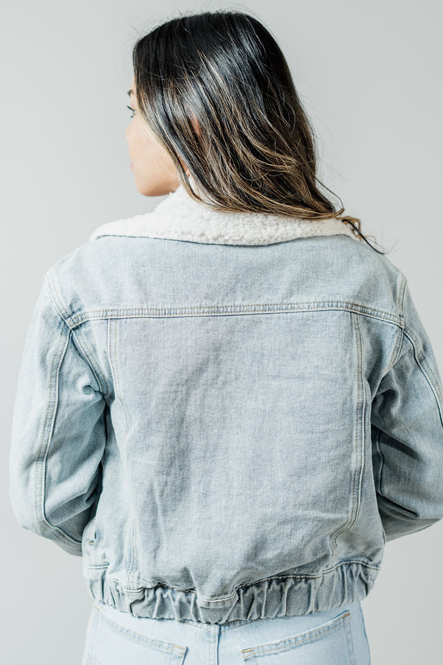 Pictured is a light-wash jean jacket with a white fuzzy collar, cuffed sleeves, button-down front, and elastic waistband.