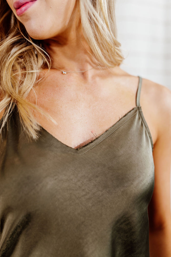 Pictured is an olive-green, satin tank top with adjustable straps, distressed neckline, and raw hemline.