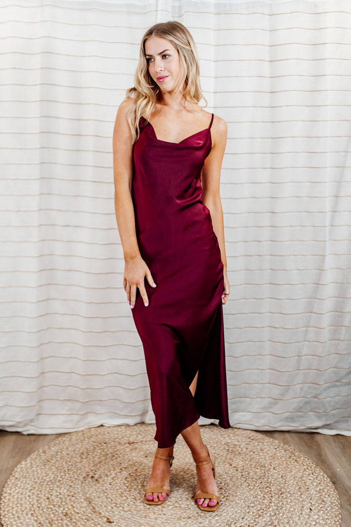 Pictured is a red, stunning and silky satin slip dress with a flattering cowl neckline, slit on the side, adjustable straps, and midi length.