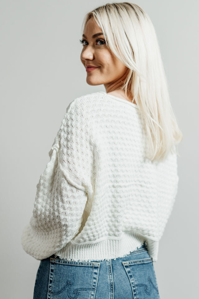 Pictured is a white, knit sweater with a scoop neckline, cropped body, and cuffed knit sleeves.