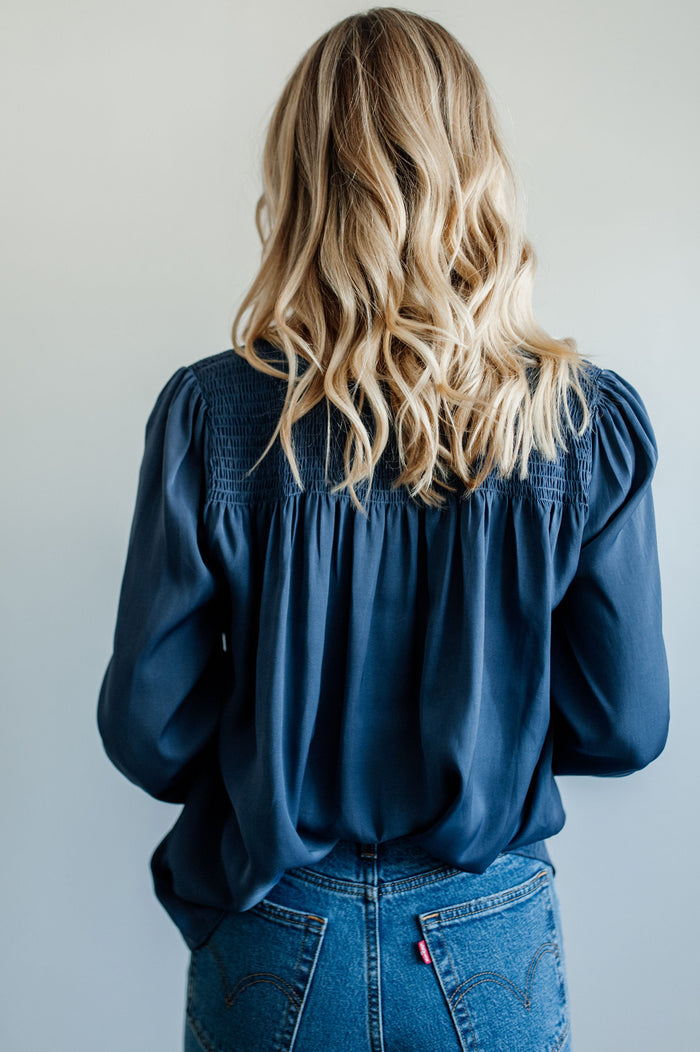 Pictured is a blue, flowy blouse with a v-neckline, balloon sleeves, and pleated back panel.