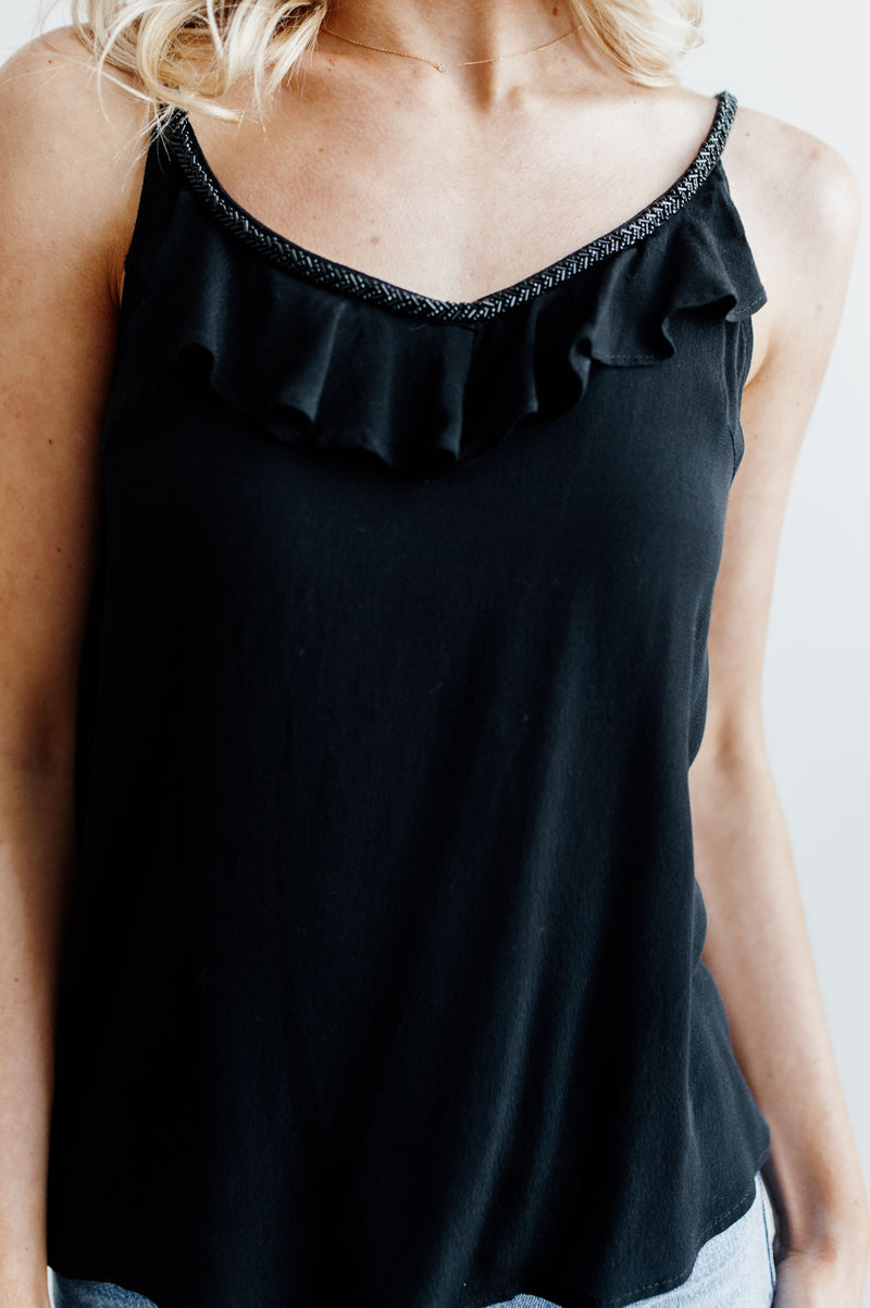 Pictured is a black, trendy top with beaded neckline, ruffle detailing, and tie back with tassels.