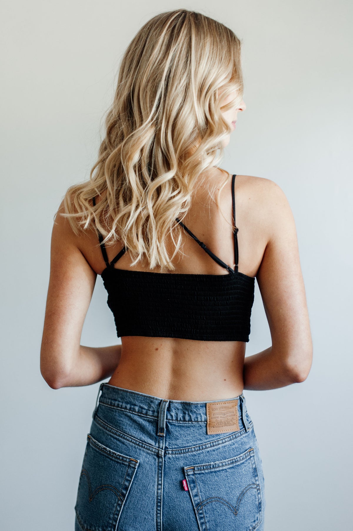 Pictured is a black, lace bralette garment with strappy criss-cross shoulder straps, ruched backing, padded cups, and cropped style.