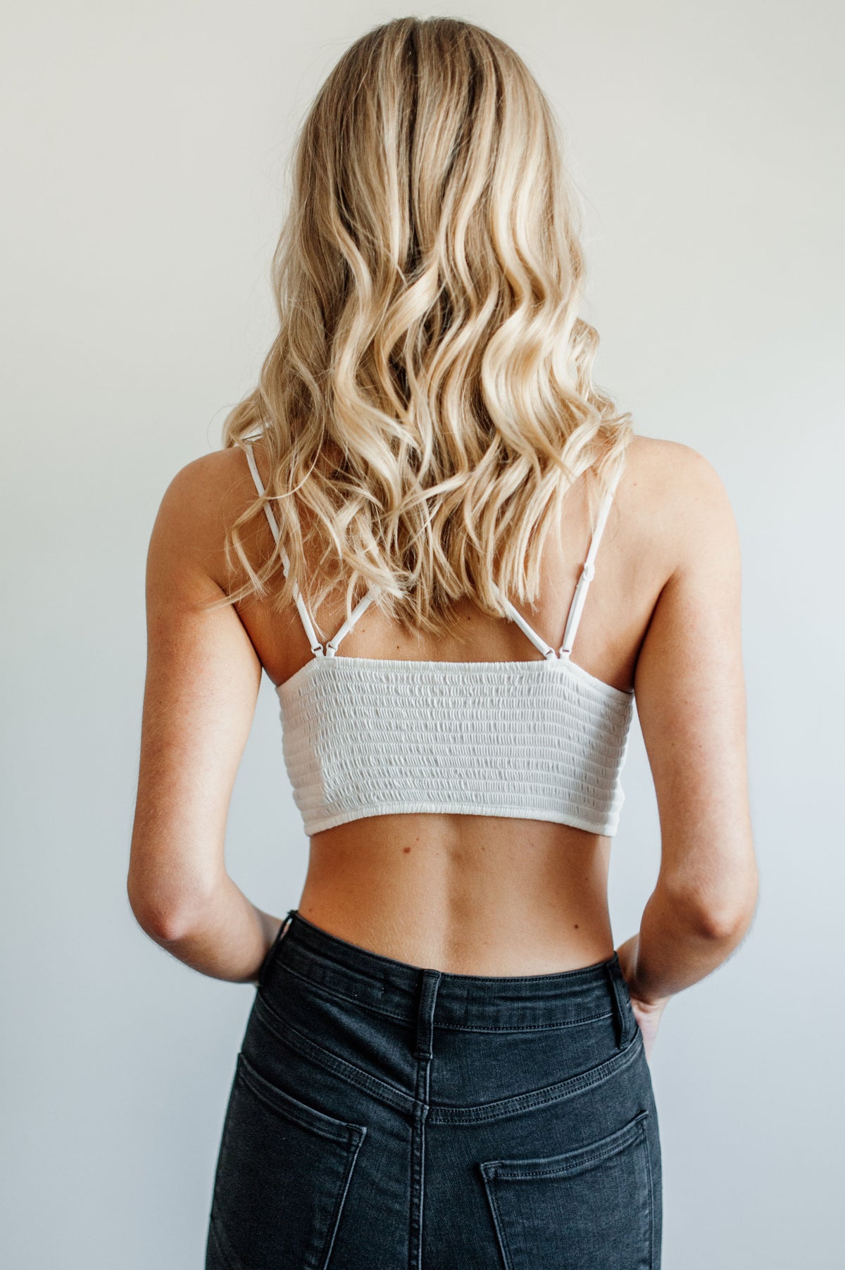 Pictured is a white, lace bralette garment with strappy criss-cross shoulder straps, ruched backing, padded cups, and cropped style.