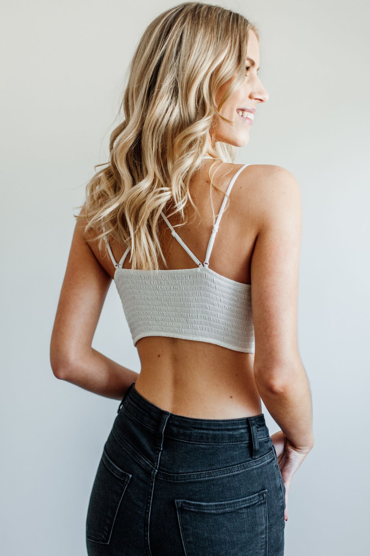 Pictured is a white, lace bralette garment with strappy criss-cross shoulder straps, ruched backing, padded cups, and cropped style.