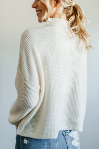 Pictured is an ivory knit sweater with a mock turtleneck, oversized body, and cuffed knit sleeves.