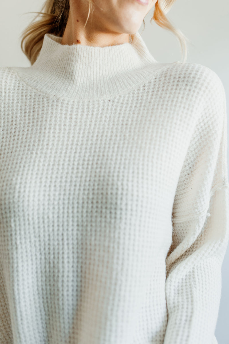 Pictured is an ivory knit sweater with a mock turtleneck, oversized body, and cuffed knit sleeves.