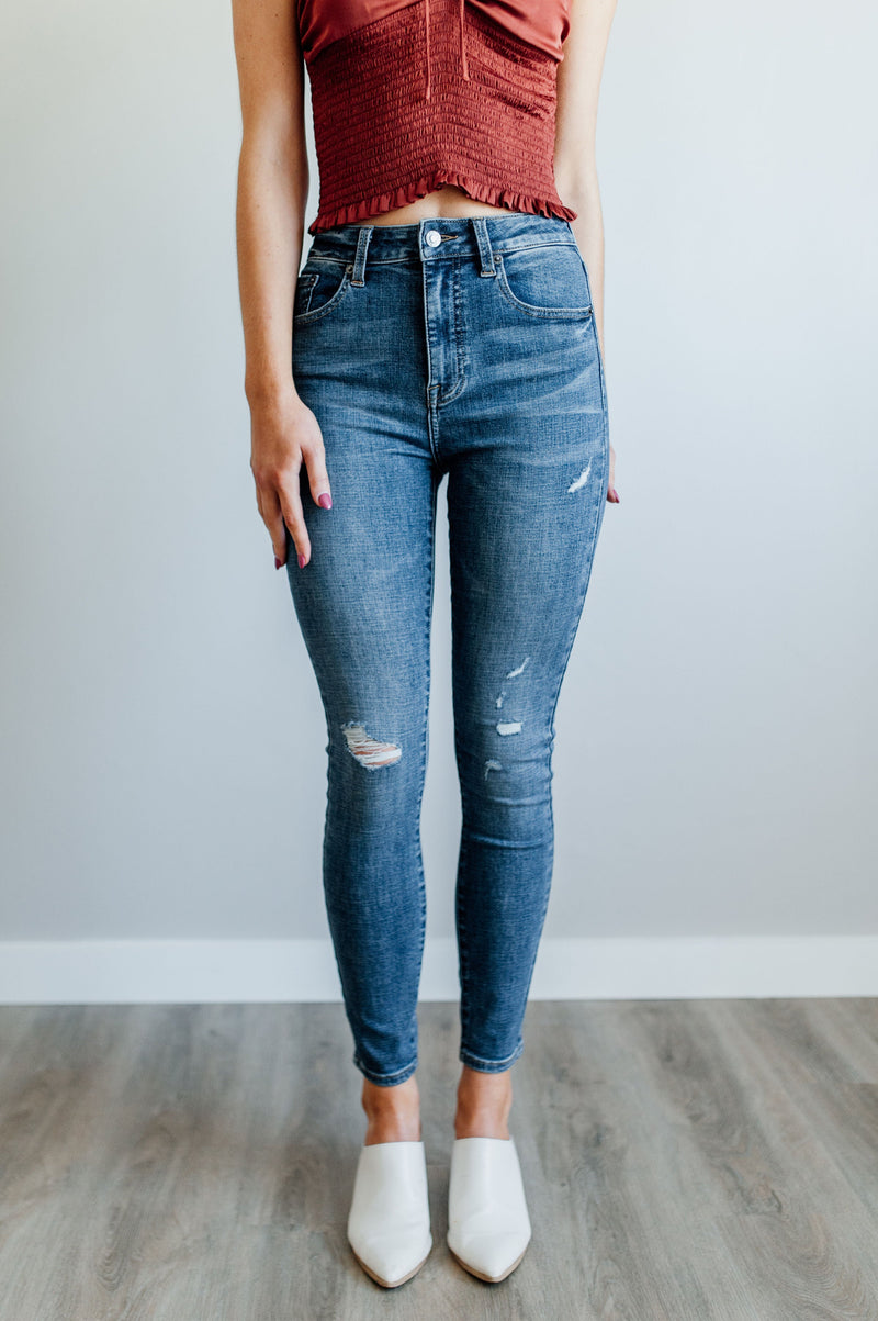 Pictured is a mid-wash, distressed denim jean with a high-rise waist, cropped ankles, and lightly distressed denim material.