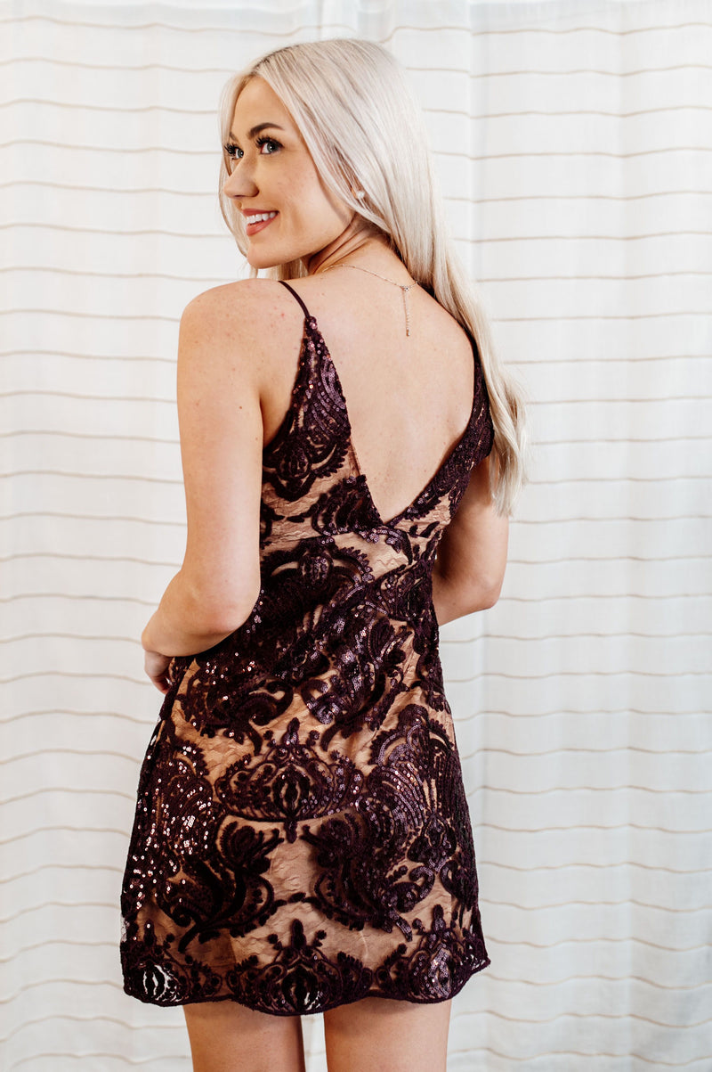 Pictured is a burgundy and nude sequin mini dress with low v-neckline, intricate lace detailing, and embroidery with added sequins. 